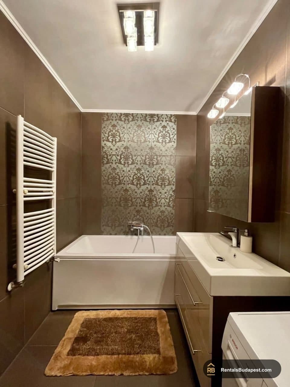Brown-tiled bathroom with white bathtub, patterned feature wall, heated towel rail and vanity sink, Sobieski János utca