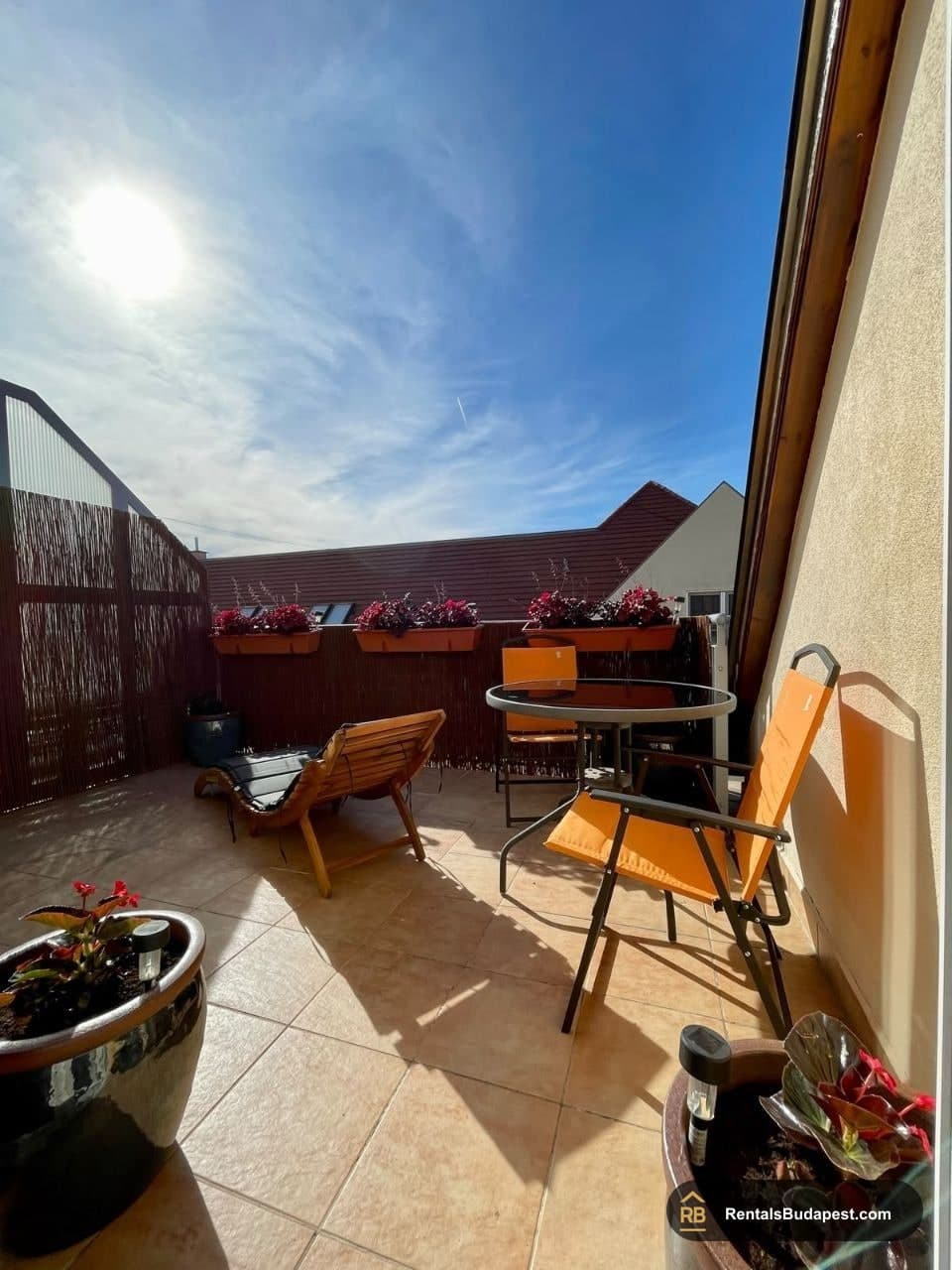Sunny tiled balcony with wooden lounger, orange folding chairs, round table and potted plants, Sobieski János utca
