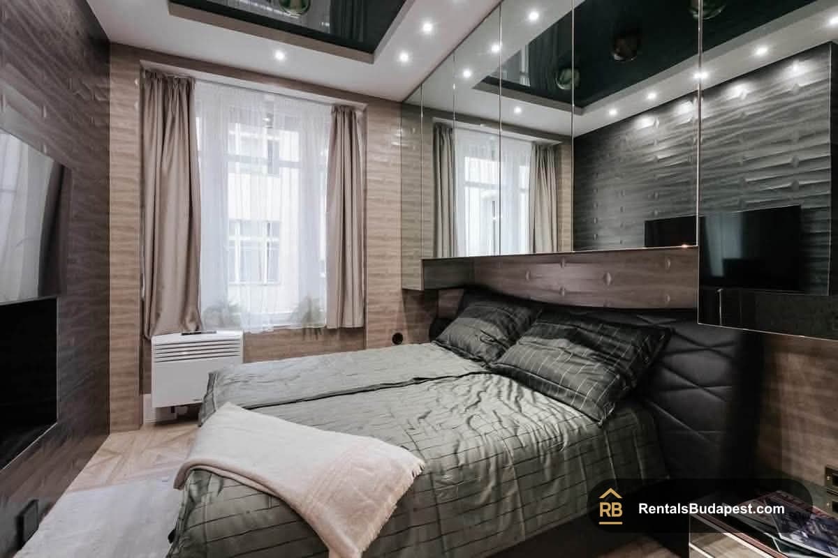 Bedroom with a made double bed, mirrored wall panels, glossy black ceiling, and curtained window with radiator