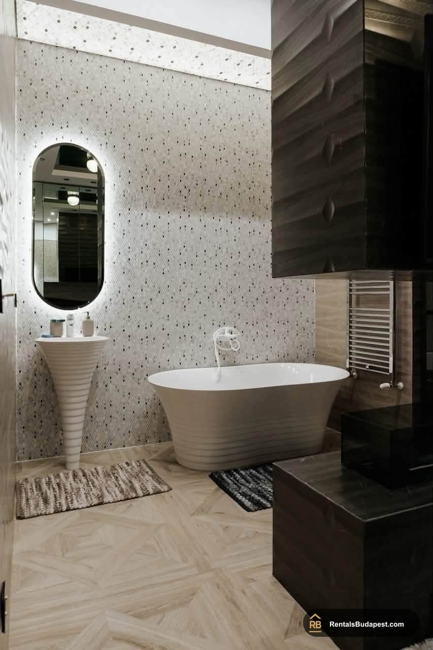 Bathroom with freestanding white bathtub, pedestal sink, backlit oval mirror, and patterned tiled wall