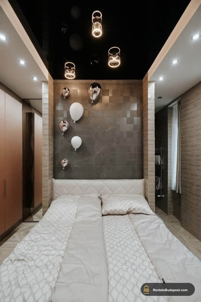 Bedroom with white quilted bed, balloon-shaped wall decor, black ceiling, and warm pendant lights above
