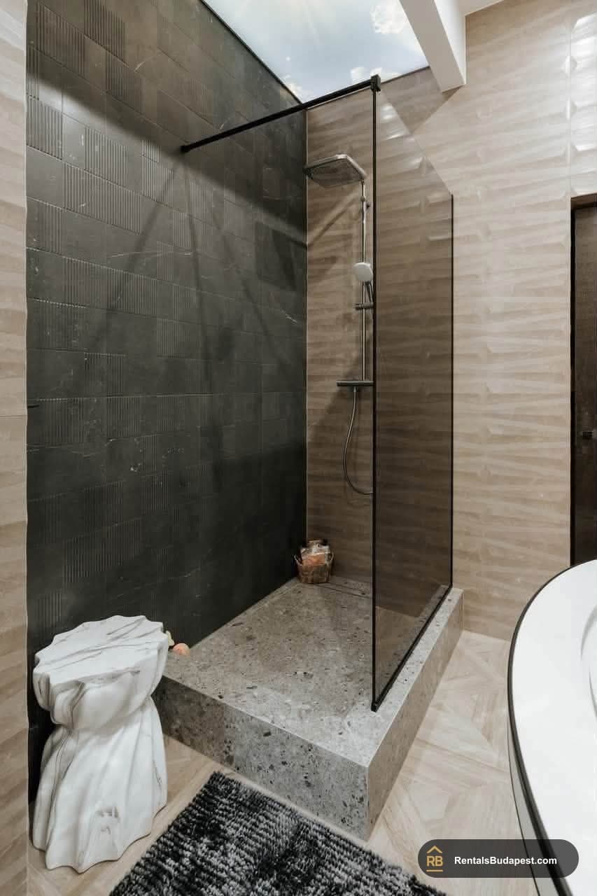 Walk-in shower with dark tiled wall, stone base, glass screen and white tub edge, Akadémia utca