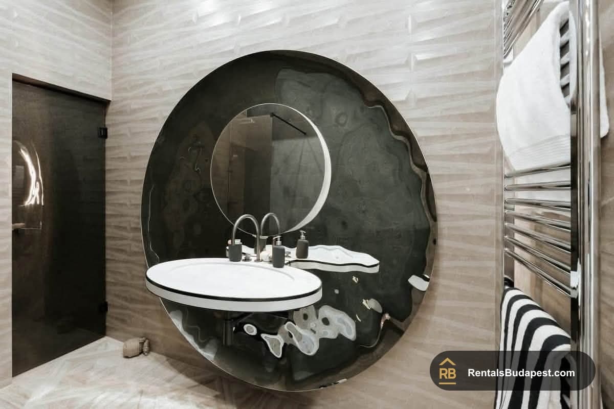 Round mirror above white wall-mounted sink with chrome towel warmer and striped towel, Akadémia utca