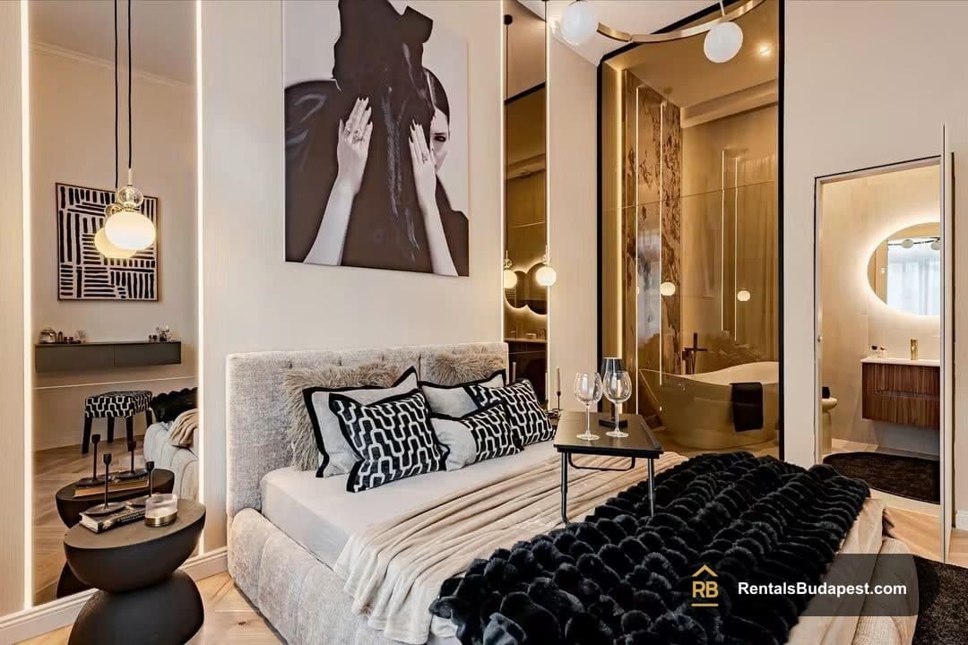 Bedroom with grey upholstered bed, monochrome patterned pillows, black faux-fur throthrow, and glass-walled bathroom with freestanding tub, Falk Miksa utca