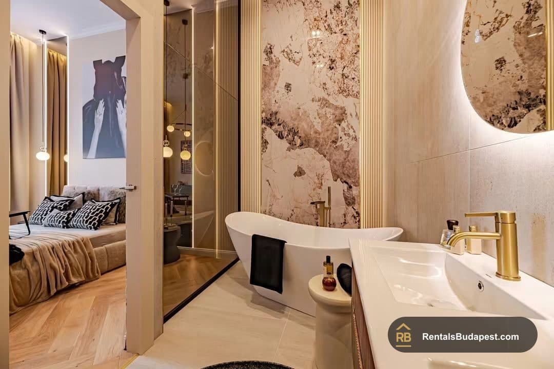 Bathroom with freestanding white tub, marble-look feature wall, gold tap, round mirror, and view through doorway into bedroom, Falk Miksa utca