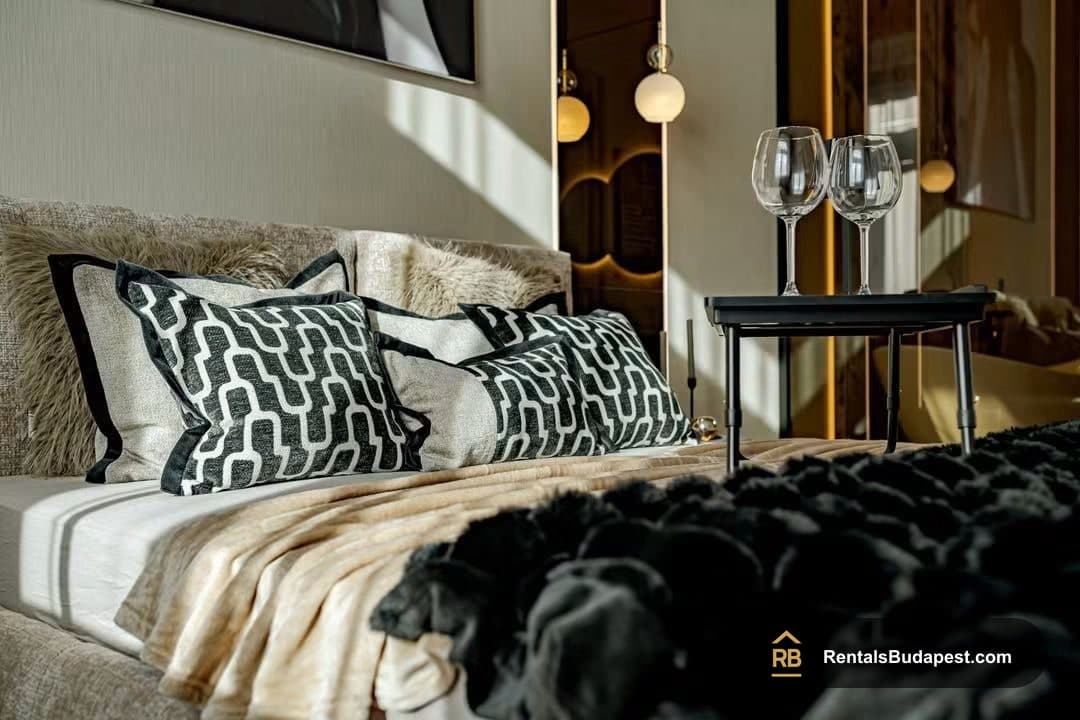 Close view of bed with patterned black and white cushions, black faux-fur blanket, and two wine glasses on a black side table, Falk Miksa utca