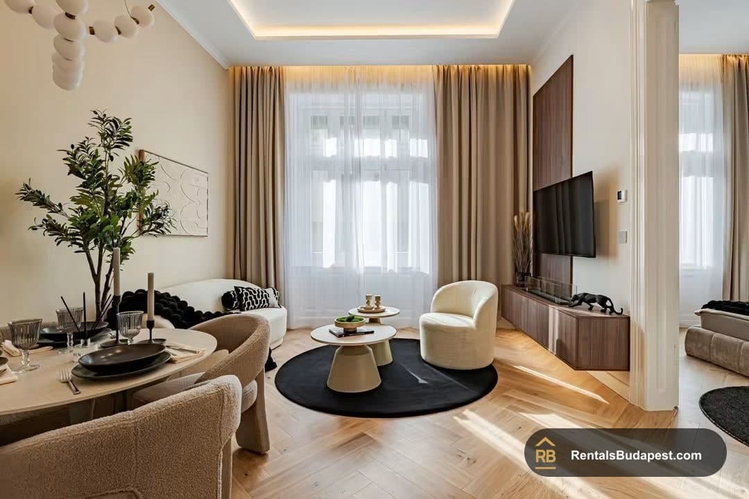 Living room with dining table set for two, cream armchair, round black rug, wall-mounted TV, and tall windows with sheer curtains, Falk Miksa utca
