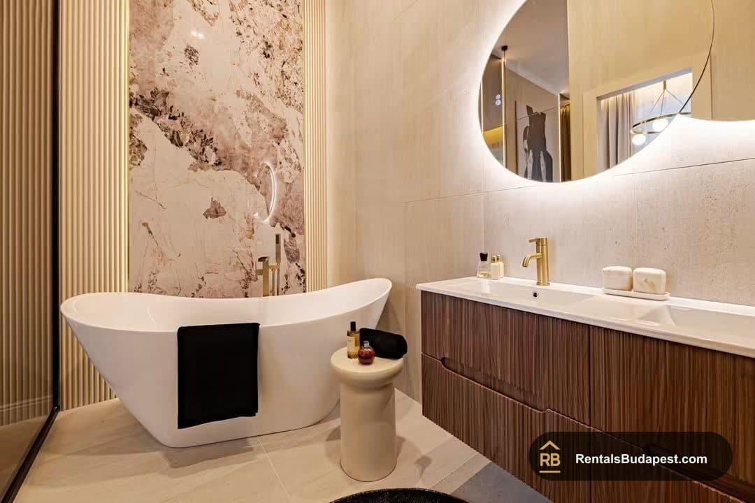 Bathroom with white freestanding tub beside marble-look wall, wood-finish vanity with gold tap, and backlit round mirror, Falk Miksa utca