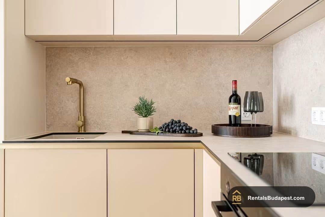 Kitchen with beige cabinets, gold tap over inset sink, bottle of red wine, grapes on a board, and induction hob, Falk Miksa utca