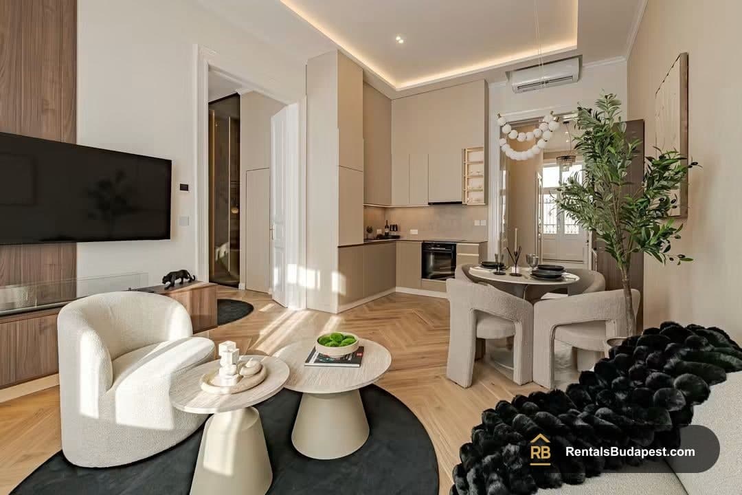 Open-plan living room with wall TV, cream armchair, round coffee tables, herringbone floor, and kitchen with dining set beyond, Falk Miksa utca