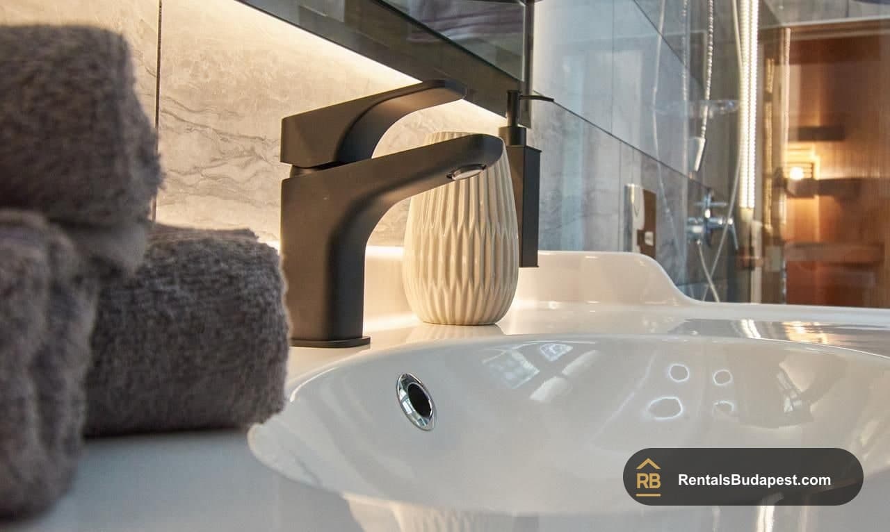 Close view of black tap and white ceramic vase on a basin beside grey folded towels, Szent István körút