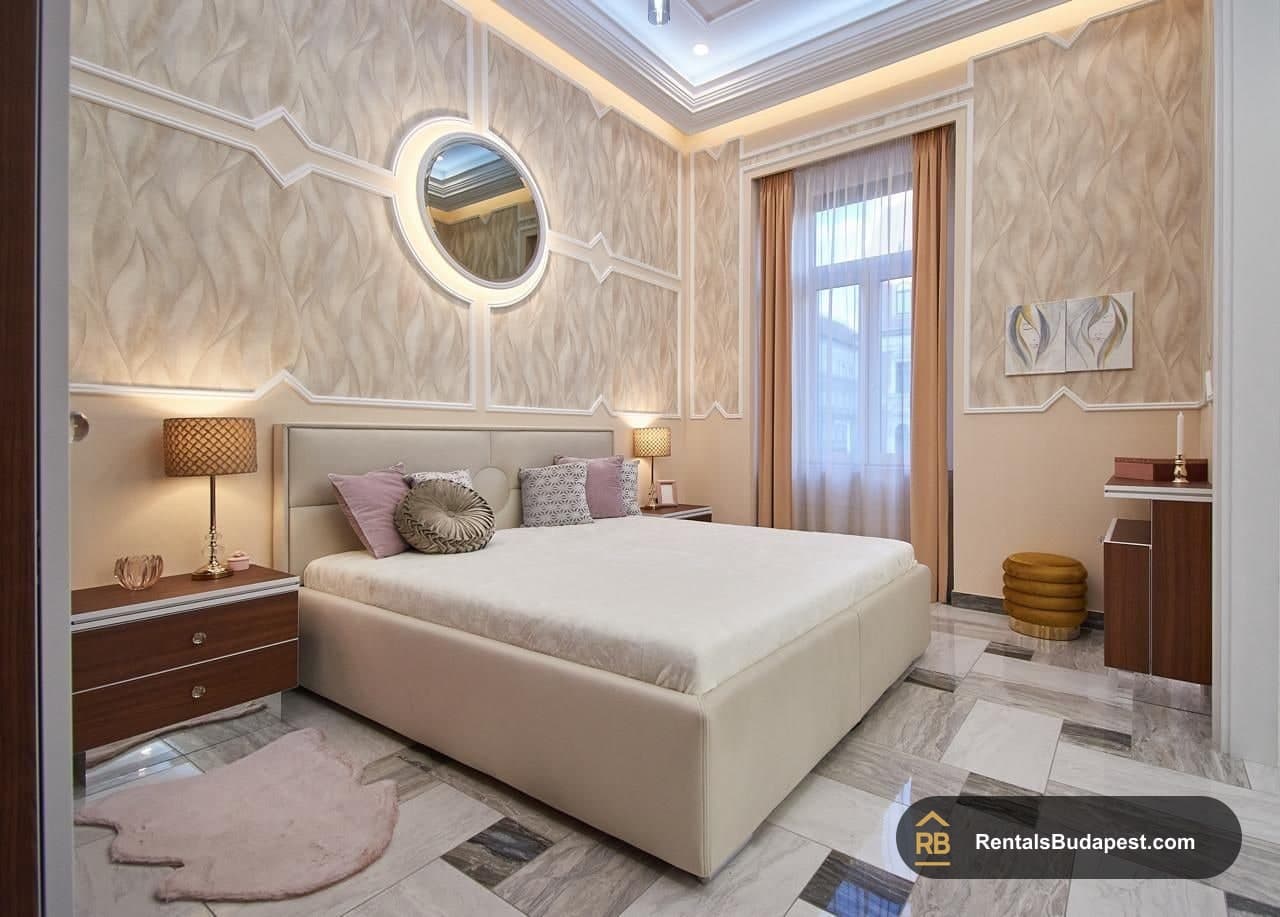 Bedroom with cream upholstered bed, pink cushions, round mirror and marble floor