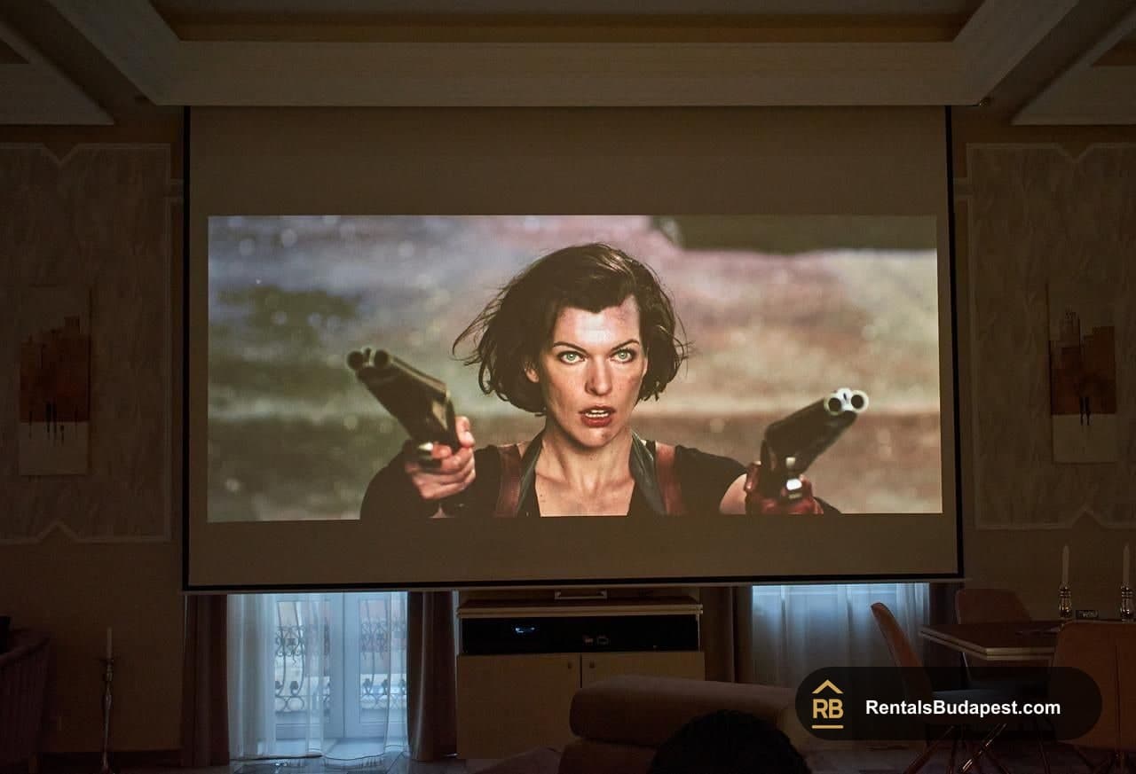 Wall-mounted projector screen showing an action film scene with a woman aiming two pistols