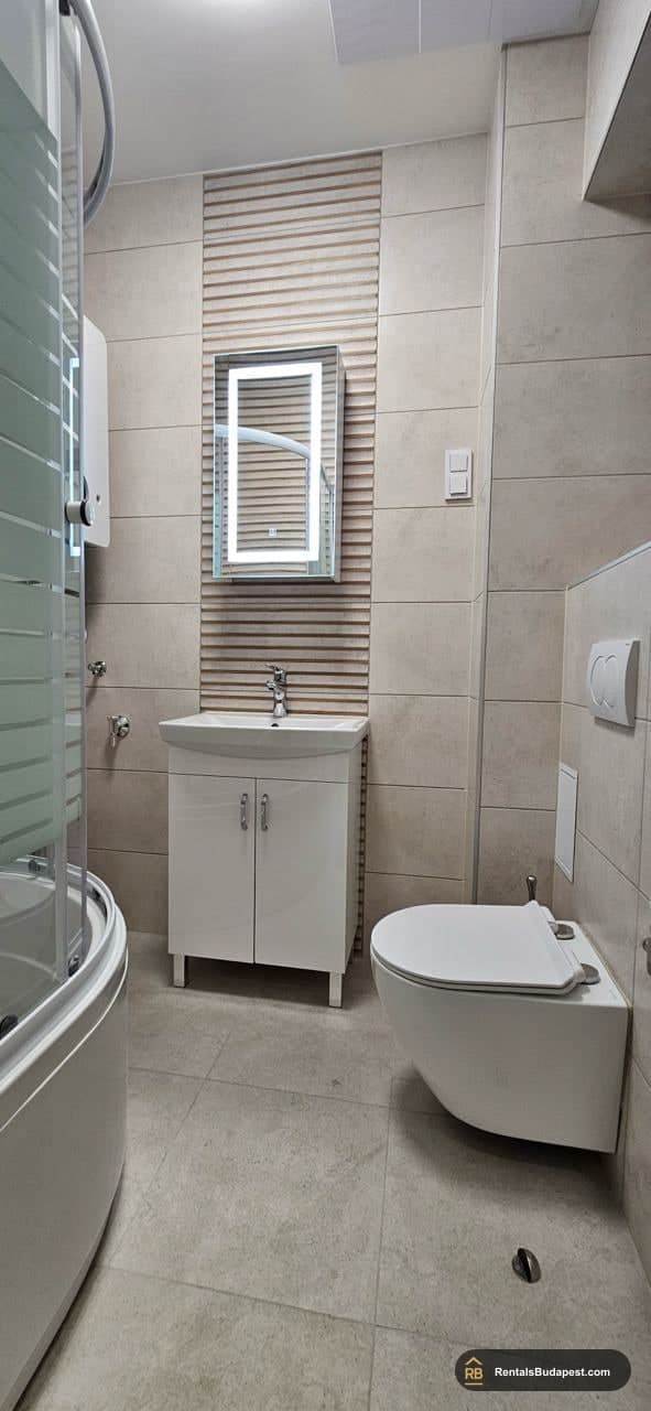 Bathroom with corner shower, wall-hung toilet, and white vanity sink under a backlit mirror