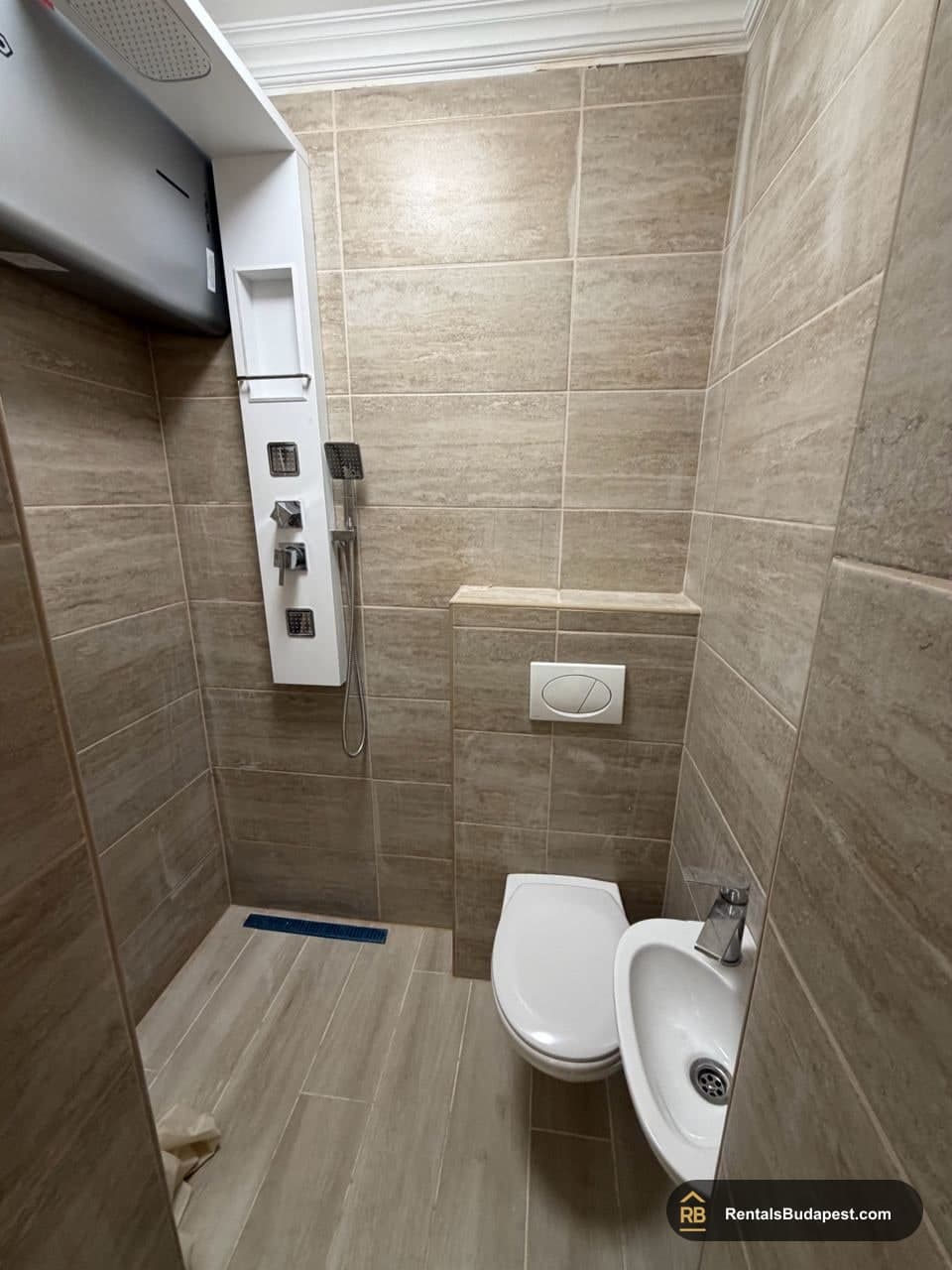 Compact bathroom with beige tiled walls, wall-mounted toilet, corner shower with handheld spray and small sink, Szent István körút
