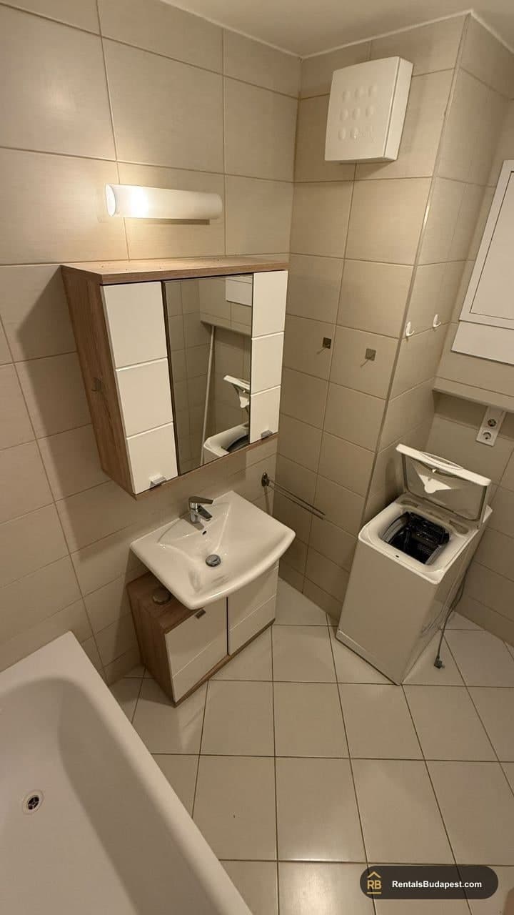 Bathroom with white sink and wood-effect cabinet, washing machine beside a bathtub, beige tiled walls, Viola utca