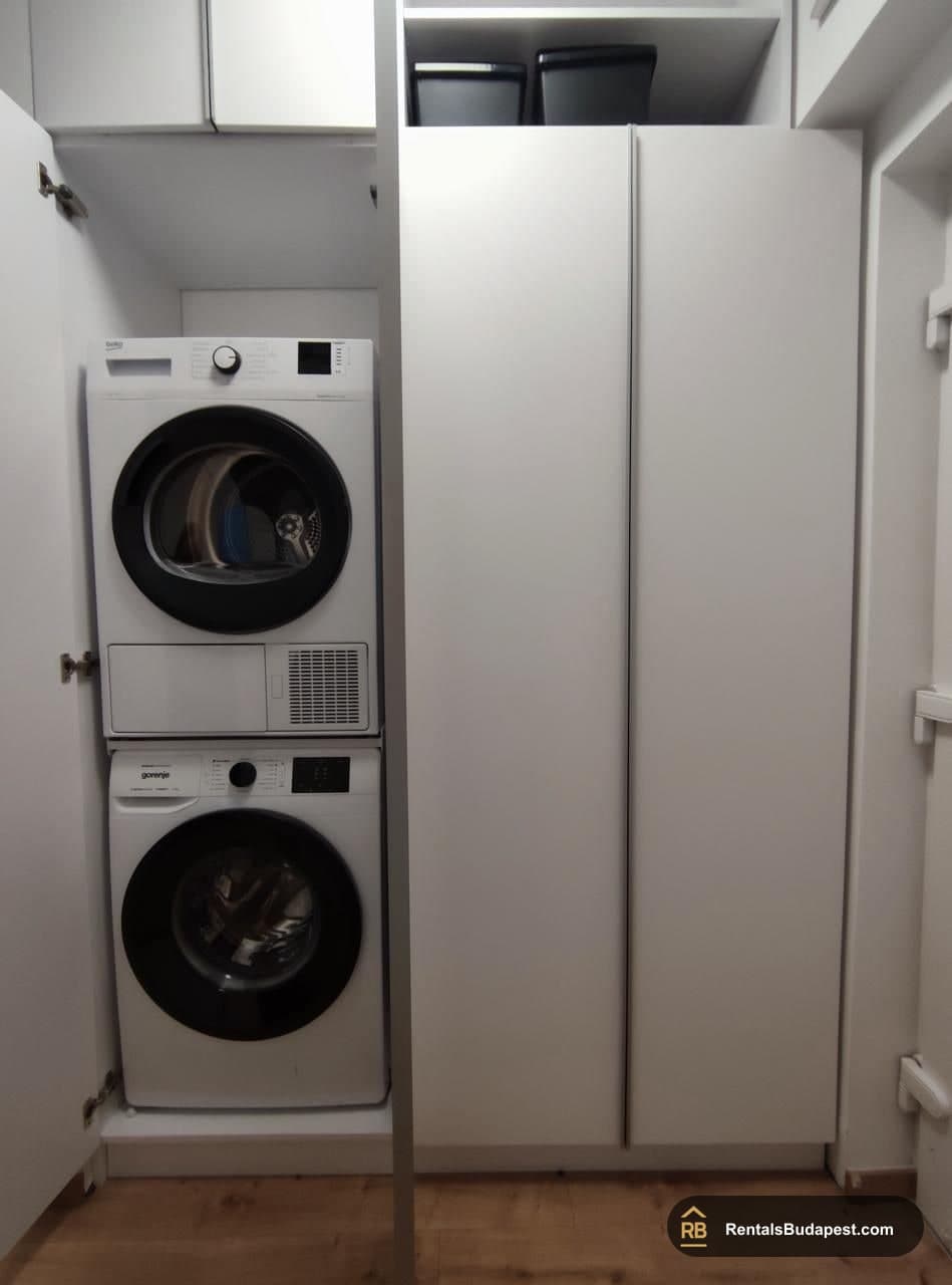 Stacked white washing machine and tumble dryer set inside a tall white cabinet with closed doors, Corvin sétány