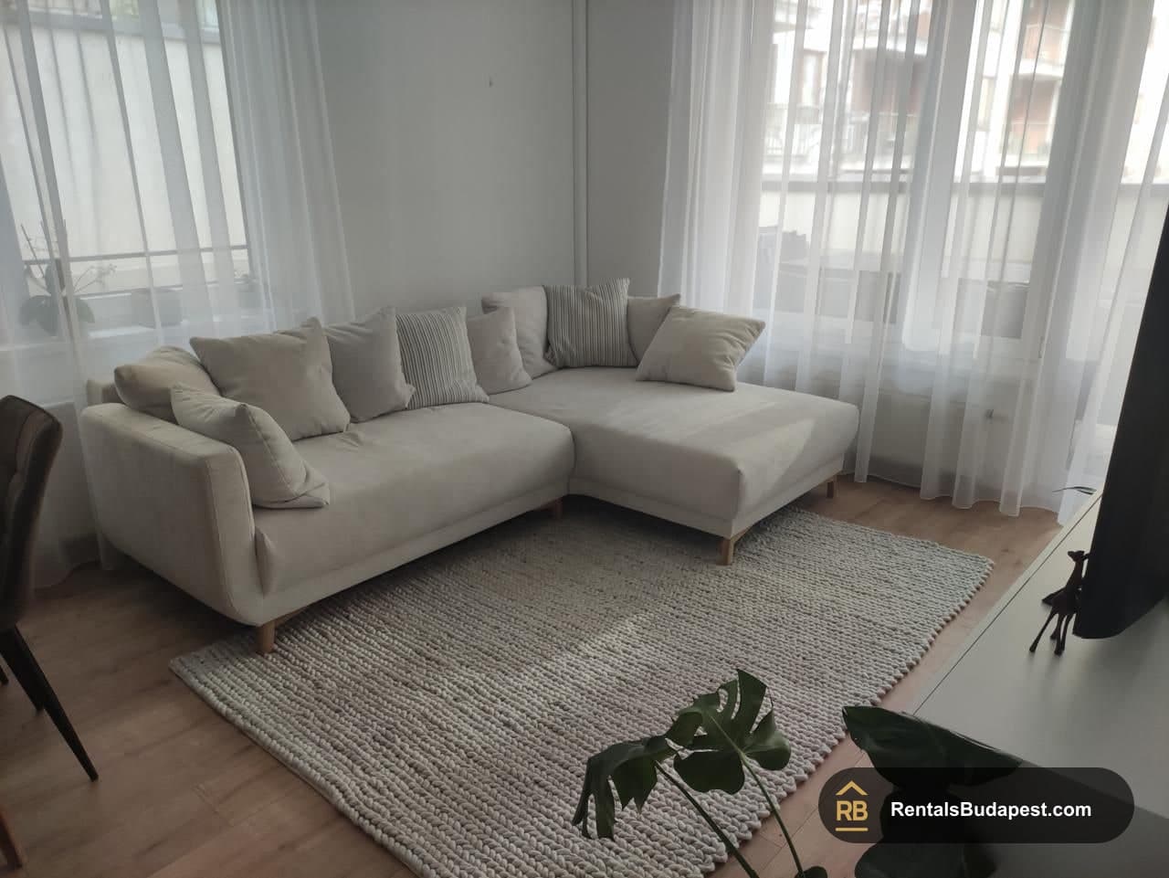 Cream corner sofa with striped cushions on a textured rug beside sheer curtained windows, Corvin sétány