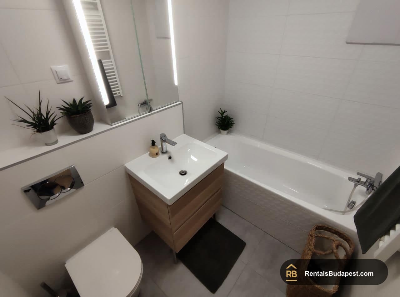 White bathroom with wood-effect vanity, bathtub, wall-mounted toilet and backlit mirror, Corvin sétány