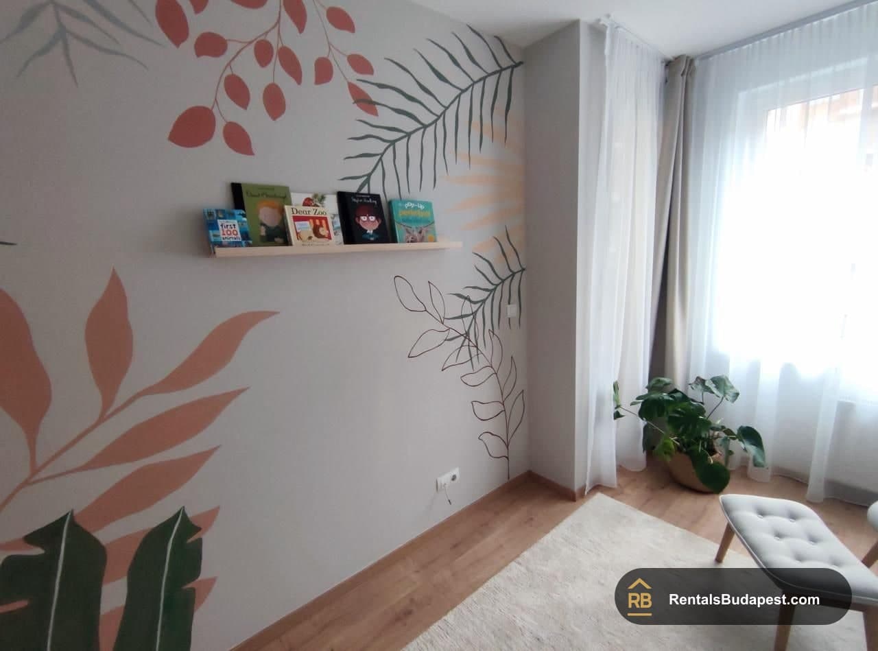Child's bedroom with hand-painted tropical leaf murals, a wall bookshelf holding picture books, and sheer curtains by the window, Corvin sétány