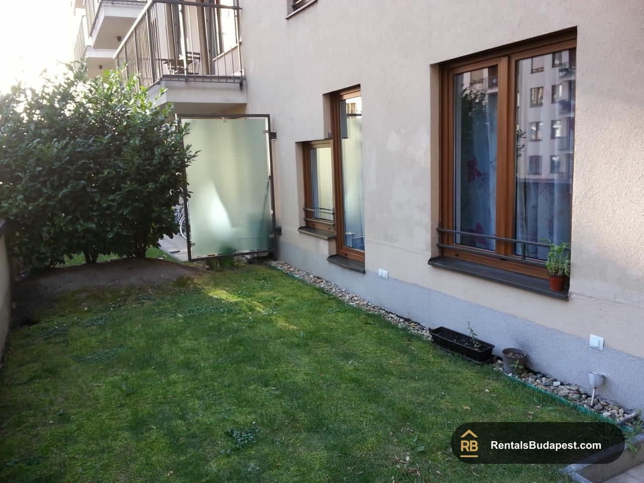 Private back garden with a lawn, shrubs, gravel border, and ground-floor windows of the building, Corvin sétány