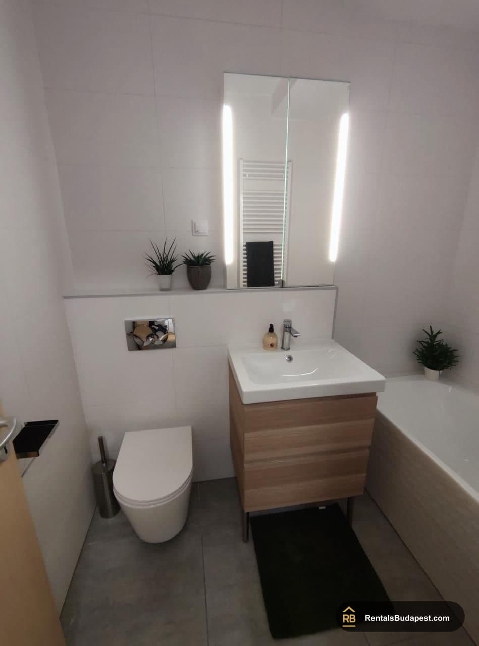 Bathroom with a wall-hung toilet, wood-effect vanity with white basin, illuminated mirror, and a bathtub, Corvin sétány