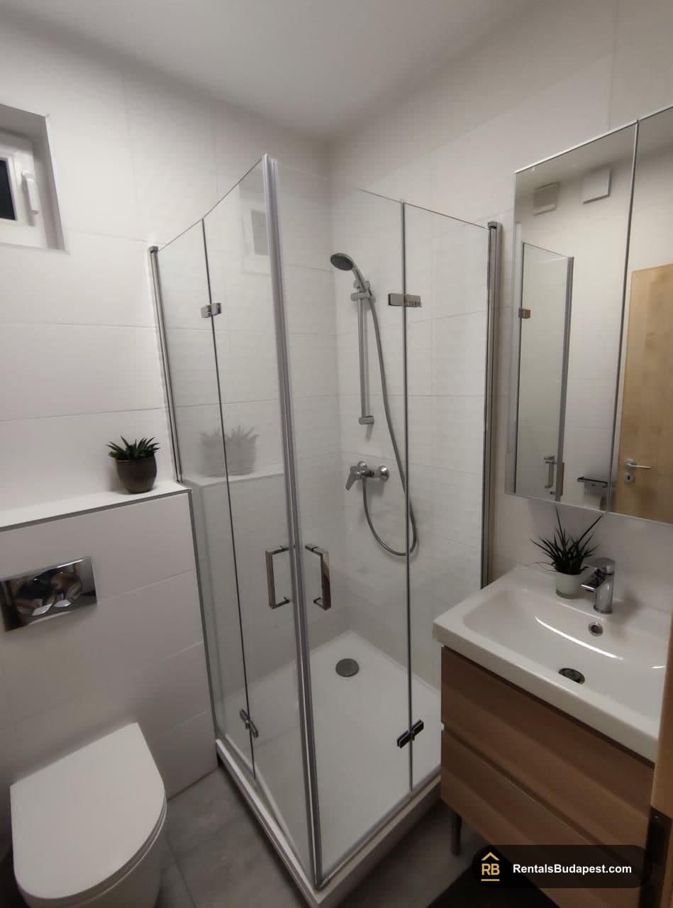 Bathroom with a corner glass shower cubicle, white tiled walls, wood-effect vanity, and a small window, Corvin sétány