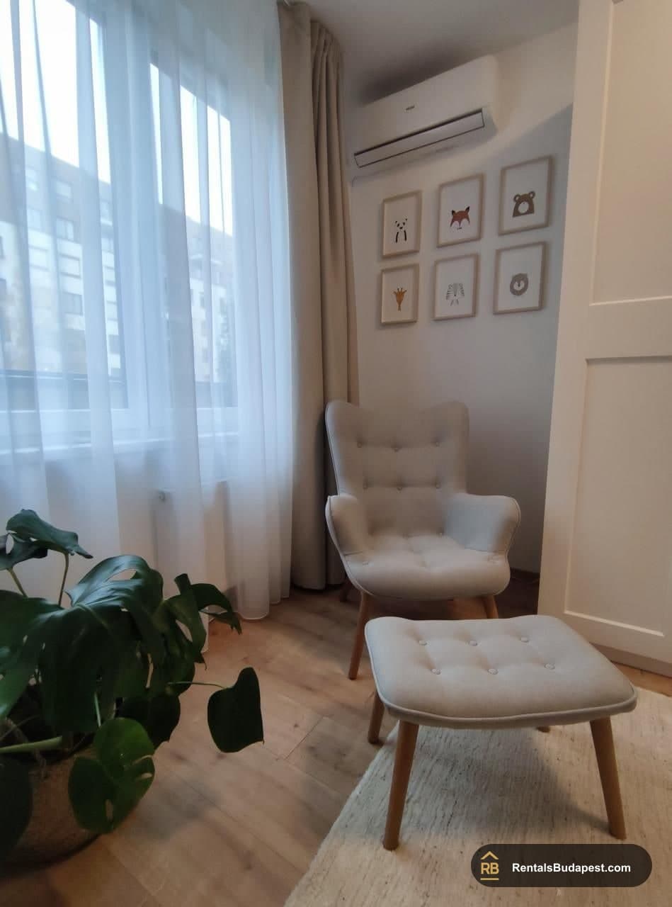 Living room corner with sheer curtains, a wall air conditioner, two light armchairs with a footstool, and framed animal prints, Corvin sétány