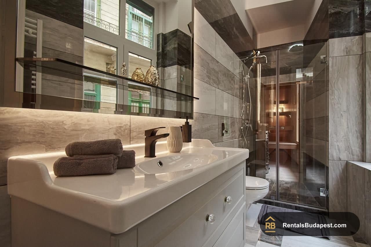 Bathroom with grey marble-look tiles, white vanity with brown tap and folded grey towels, glass shower enclosure, Szent István körút